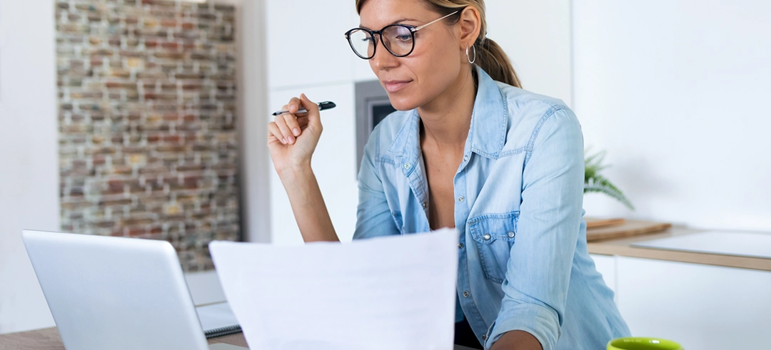 Chica comparando unos documentos mientras mira su ordenador portátil