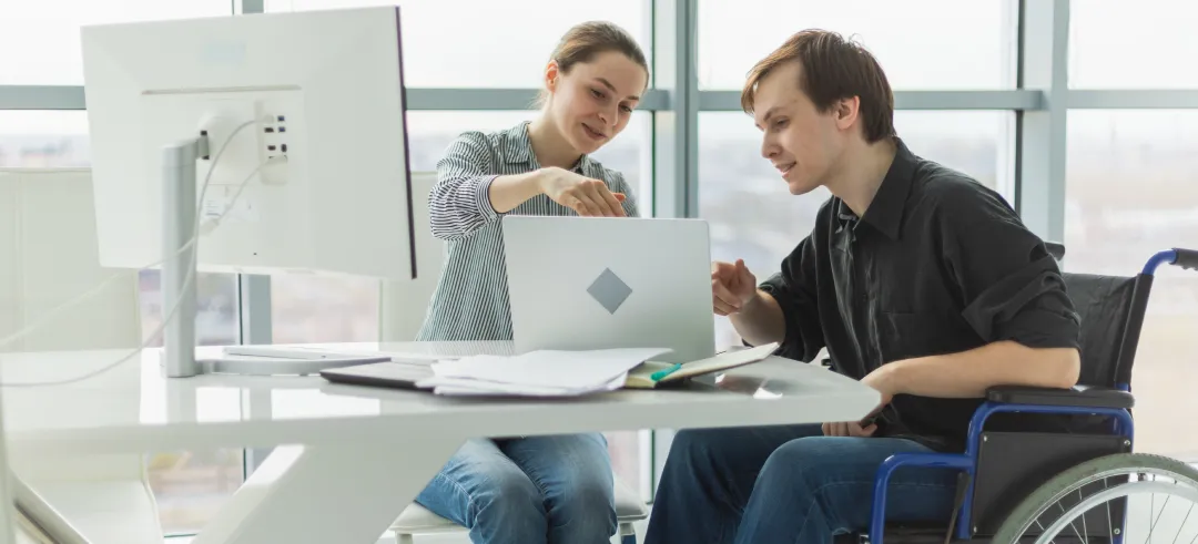 Dos personas trabajando juntas frente a un portátil en una oficina, una de ellas tiene una silla de ruedas